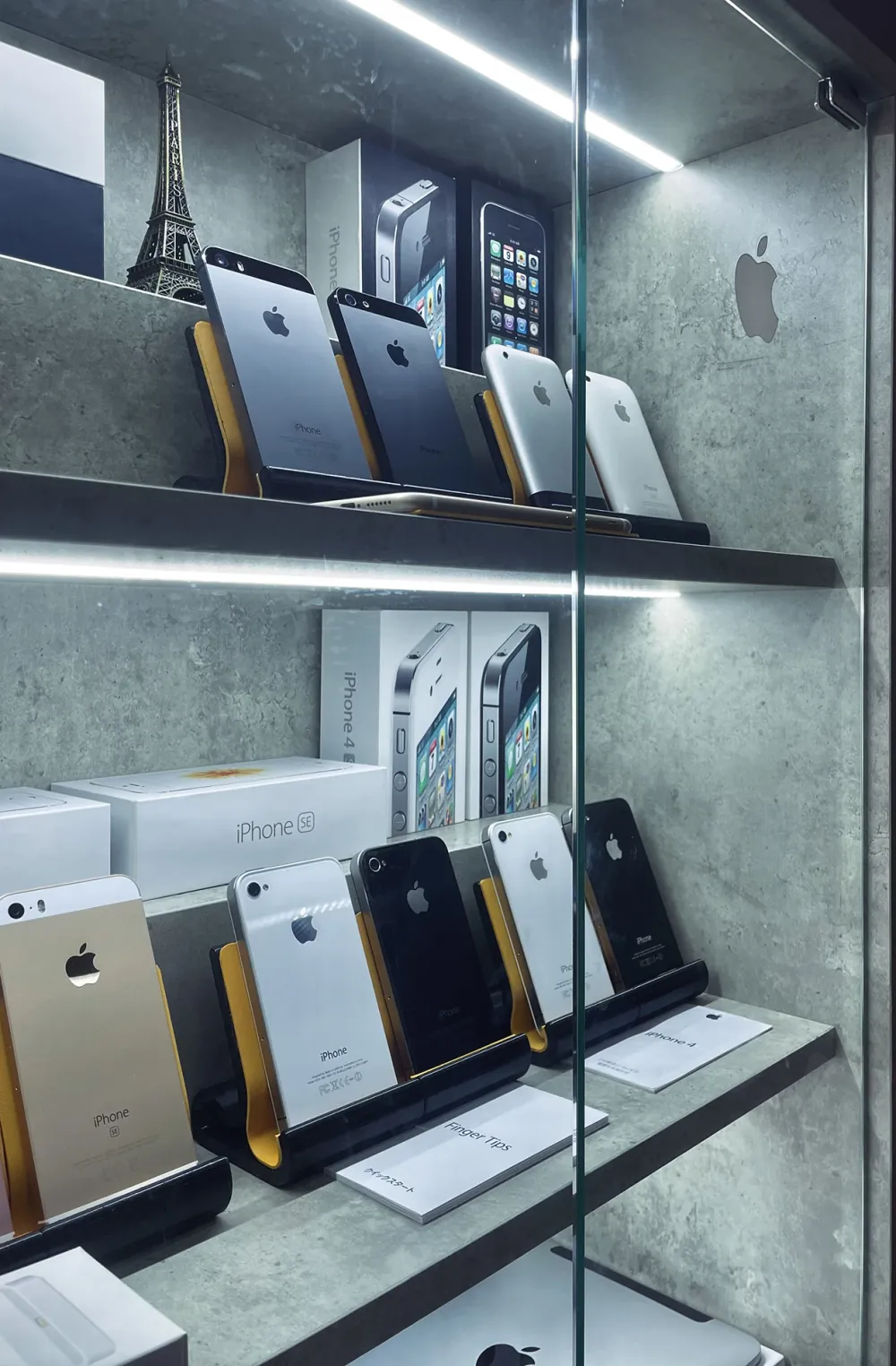 Vintage iPhones on stands across two lit shelves, seen at an angle through the glass door, with their boxes behind them.