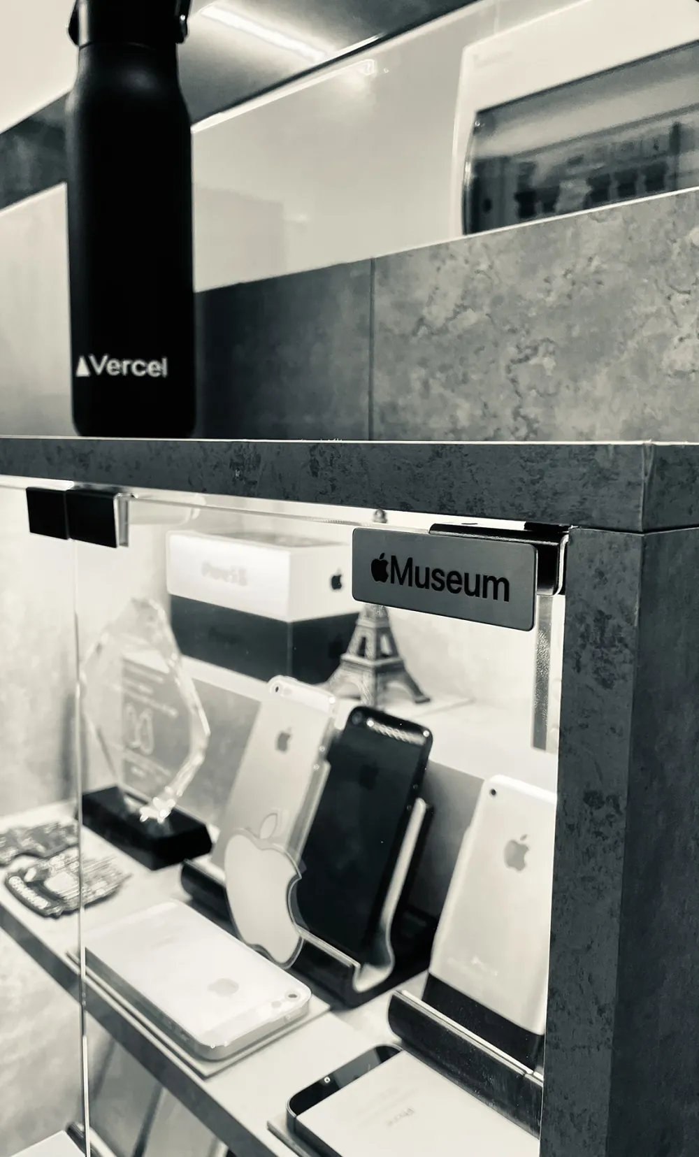 A black-and-white close-up of the Apple Museum sign on the cabinet door, with a Vercel bottle on the shelf above.