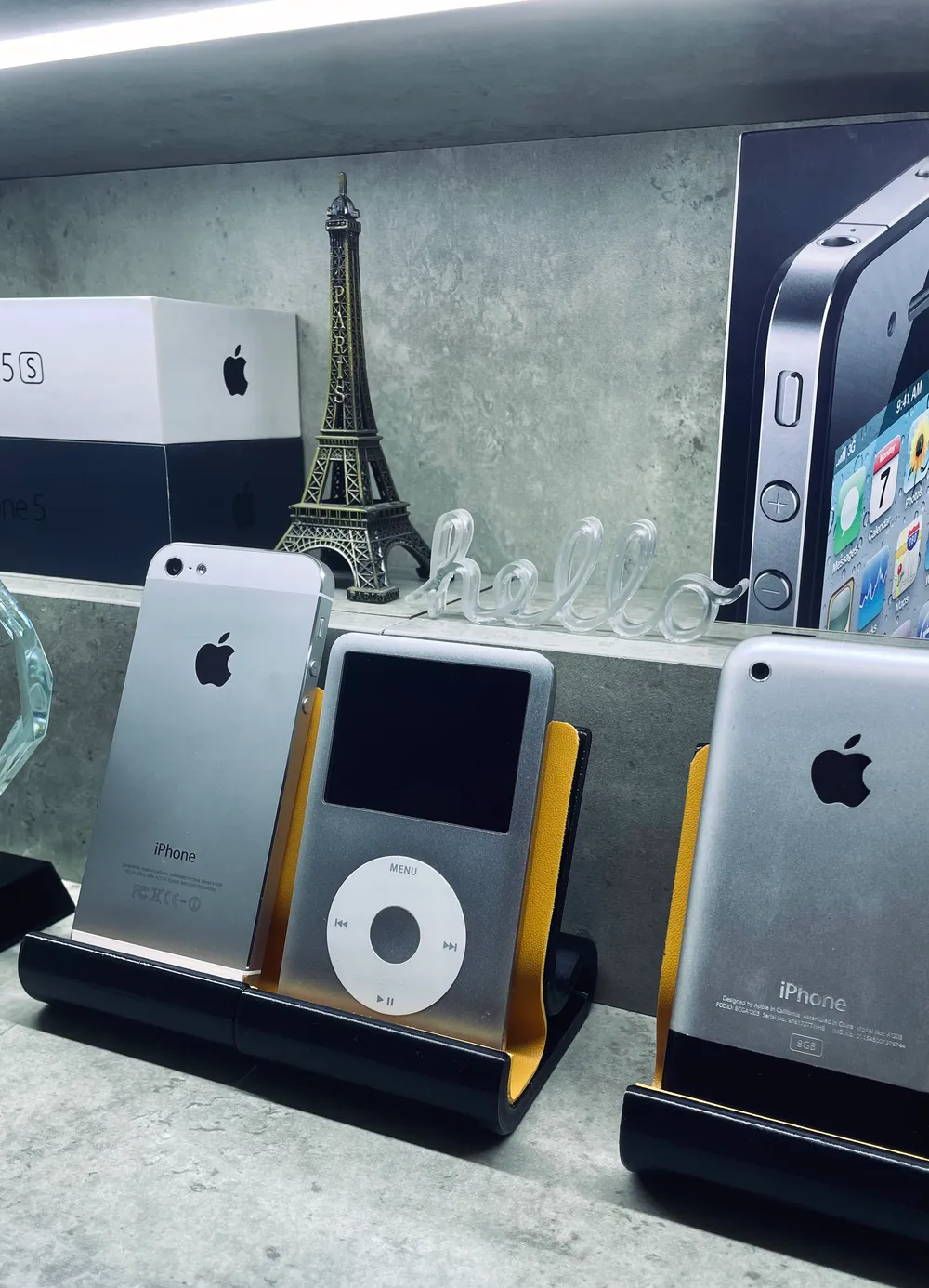 A silver iPhone 5, an iPod classic, and the original iPhone on black stands, below an iPhone 5s box, an Eiffel Tower model, and a glass hello sign.